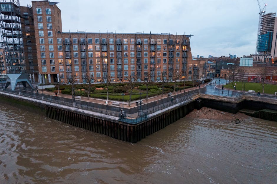 A narrow outdoor pathway alongside a modern brick building with large glass windows, part of the Blackfriars station area garden maintenance for flats. The garden beds on the left contain a variety of